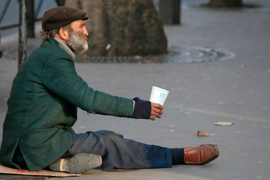 A man sits on a piece of cardboard in Paris, France with nothing but the clothes he is wearing and a cup to collect change in.